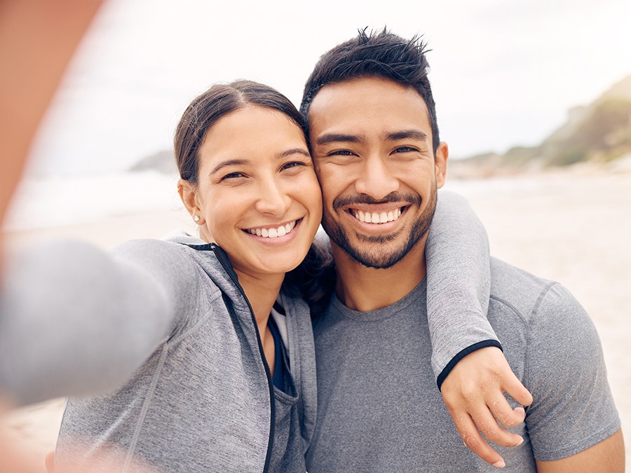 A couple standing close together on a beach, both smiling at the camera