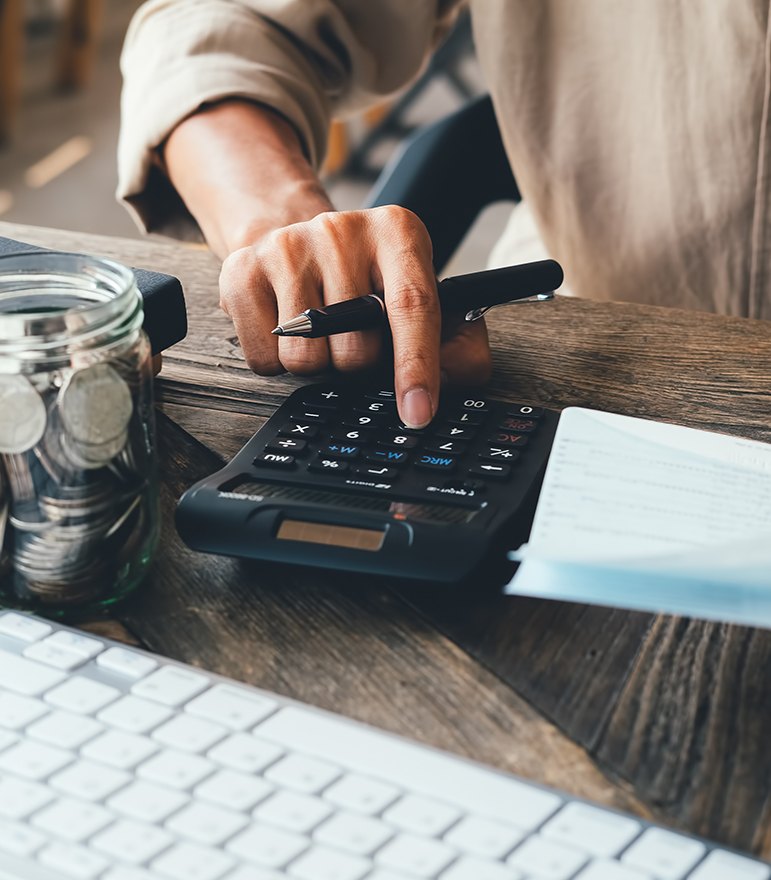 A hand using a calculator beside a jar of coins and a notebook