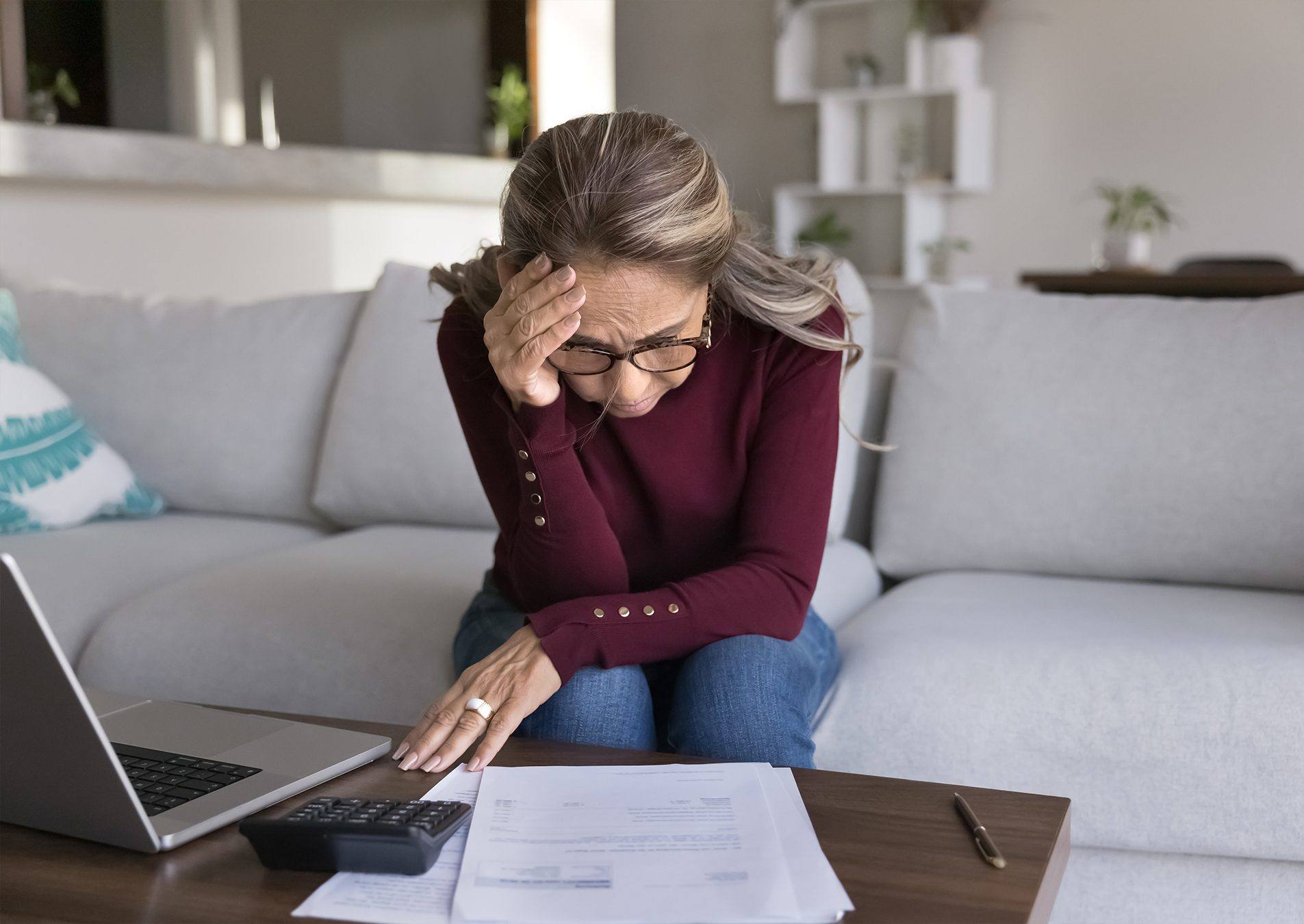 A woman at a laptop with paperwork spread out, holding her head in her hands