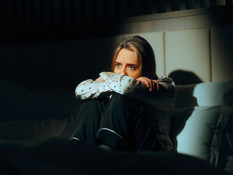 A woman sitting on a bed in a darkened room, knees drawn up to her chest